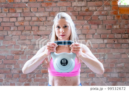 Blonde young muscular woman with sporty looks doing high pull exercise with kettlebells on hard Blonde young muscular woman with sporty looks doing high pull exercise with kettlebells on hard 99532672
