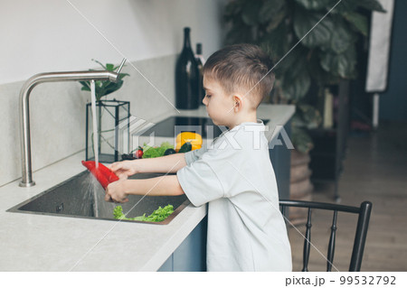 Six-year-old boy washing vegetables in the kitchen sink. Side view. Six-year-old boy washing vegetables in the kitchen sink. Side view. 99532792