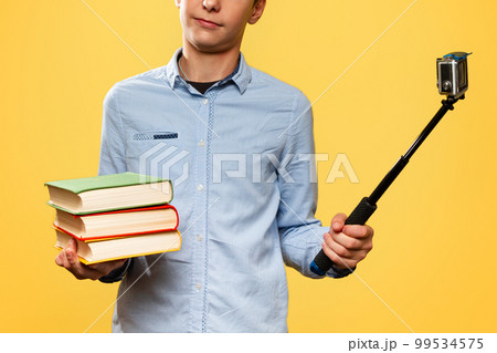 The concept of blogging and education. A disgruntled teenage boy in a blue shirt holds textbooks and takes a selfie or video on a camera with a monopod. Yellow background. Close up The concept of blogging and education. A disgruntled teenage boy in a blue shirt holds textbooks and takes a selfie or video on a camera with a monopod. Yellow background. Close up 99534575