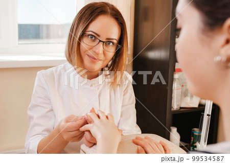 Medicine, cosmetology and manicure. A woman manicure in glasses, a mask and a white coat does a manicure to a client in the salon. View from over the visitor's shoulder. Close up 99534587