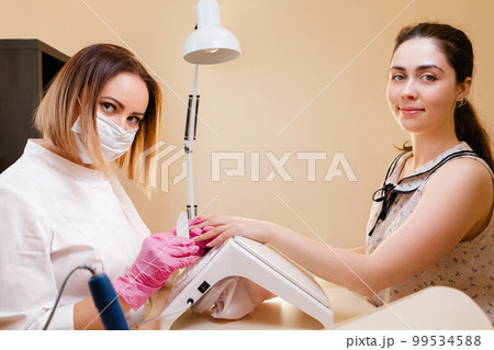 Medicine, cosmetology and manicure. A woman manicure master, in a mask and a white coat does a manicure to the client in the salon, filing her nails. Side view Medicine, cosmetology and manicure. A woman manicure master, in a mask and a white coat does a manicure to the client in the salon, filing her nails. Side view 99534588