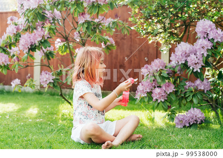 Cute little girl with toned red hair blowing soap bubbles while sitting on a green lawn in the garden. Having fun during school holidays at home. Offline activity for kids. Happy childhood concept Cute little girl with toned red hair blowing soap bubbles while sitting on a green lawn in the garden. Having fun during school holidays at home. Offline activity for kids. Happy childhood concept 99538000