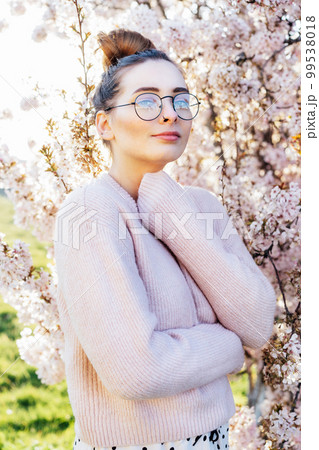 Portrait of young caucasian brunette hipster girl in glasses and light pink clothes near blooming tree with sunset light and sunbeams. Generation Z girl enjoy spring mood. Vertical card. 99538018