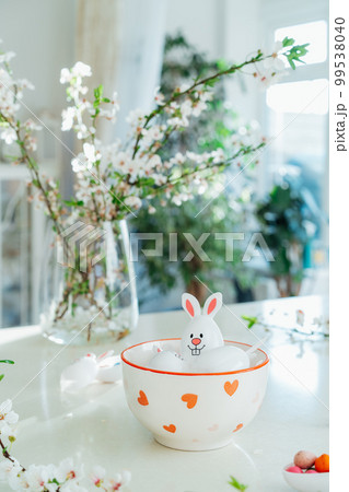 Funny Easter eggs containers with bunny ears filled with candy and chocolate in bowl on white kitchen table with blooming branches. Festive spring Easter composition. Vertical card. Selective focus 99538040