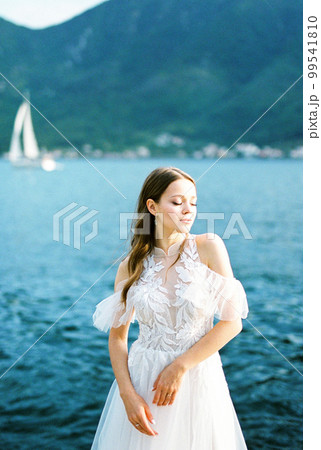 Bride in a white lace dress stands against the backdrop of the sea and mountains 99541810