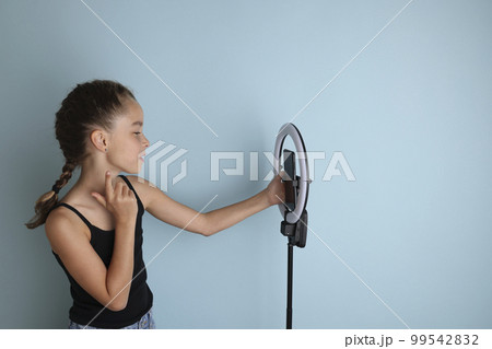 Little emotional teenage girl in a black t-shirt on an isolated blue background. Studio portrait of a young smiling teenage girl, vlogger, with a ring flash lamp and a smartphone. Girl takes a selfie. Little emotional teenage girl in a black t-shirt on an isolated blue background. Studio portrait of a young smiling teenage girl, vlogger, with a ring flash lamp and a smartphone. Girl takes a selfie. 99542832
