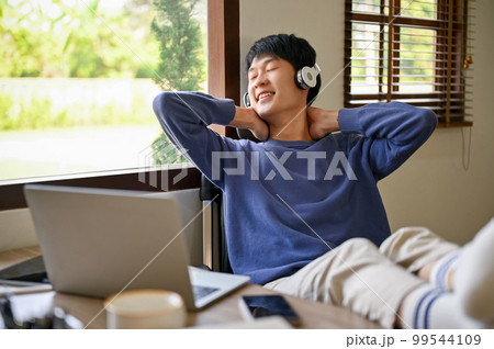 Relaxed young Asian man leaning on a chair, putting hands behind head and legs on table Relaxed young Asian man leaning on a chair, putting hands behind head and legs on table 99544109