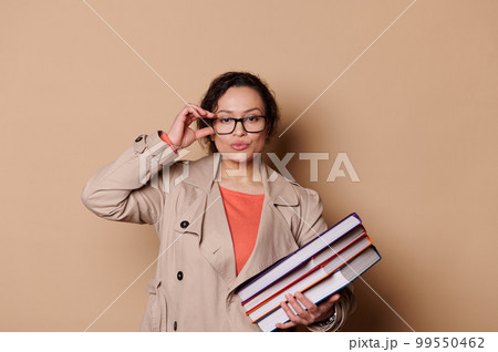 Confident middle-aged woman, wearing beige casual coat, holding a stack of hardcover books, looking at camera through her stylish spectacles, isolated on cream background. Portrait of educator teacher 99550462