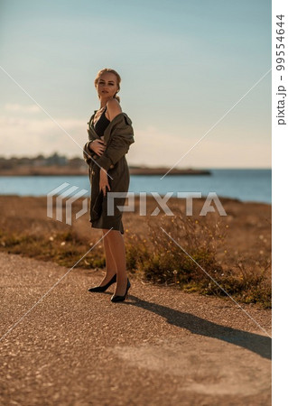 Portrait blonde sea cape. A calm young blonde in an unbuttoned khaki raincoat walks along the seashore, under a raincoat a black skirt and top 99554644