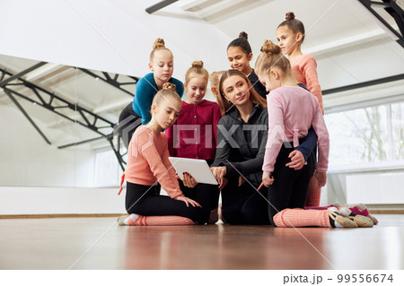 Young woman, gymnastics coach showing little girls, children video on tablet. Training session indoors. Concept of sport, childhood, rhythmic gymnastics Young woman, gymnastics coach showing little girls, children video on tablet. Training session indoors. Concept of sport, childhood, rhythmic gymnastics 99556674