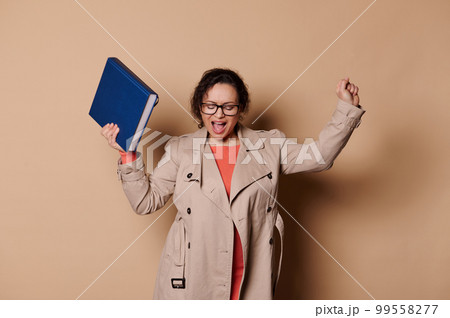 Beautiful happy female professor in casual wear and stylish eyeglasses, having fun, holding hardcover book, singing and dancing with rejoicing at the end of academic year, on cream isolated background 99558277