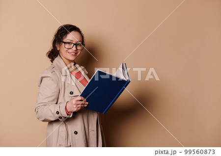 Charming middle-aged multi-ethnic woman wearing stylish spectacles and elegant beige coat, reads a book, smiles looking at camera, isolated on beige background with copy ad space 99560873
