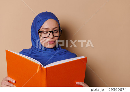 Educated sophisticated Middle-Eastern Muslim pretty woman, wearing a blue hijab and stylish eyeglasses, reading a book, isolated on a beige cream background. Erudition. Education. University concept 99560874