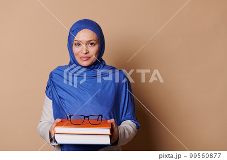 Charming Middle-Eastern Muslim woman - teacher, student, educator with head covered in blue hijab, holding pile of books and smiling confidently looking at camera, isolated on beige background 99560877