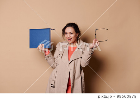 Happy positive middle-aged ethnic female teacher - educator - professor in elegant classical beige casual clothes, smiles and looks at camera while poses with a book and spectacles on cream background Happy positive middle-aged ethnic female teacher - educator - professor in elegant classical beige casual clothes, smiles and looks at camera while poses with a book and spectacles on cream background 99560878