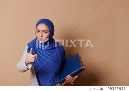 Confident strict Arab Muslim woman in blue hijab, pointing at camera, warning about the importance of reading book, standing with pile of books and looking at camera, isolated against beige background 99575931