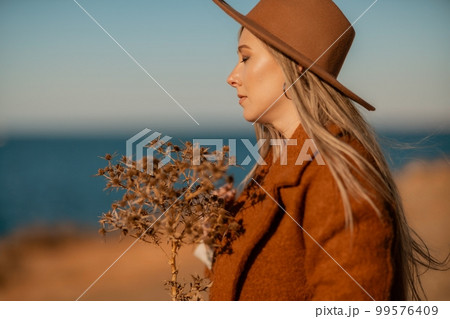 A woman walking along the coast near the sea. An elegant lady in a brown coat and a hat with fashionable makeup walks on the seashore A woman walking along the coast near the sea. An elegant lady in a brown coat and a hat with fashionable makeup walks on the seashore 99576409