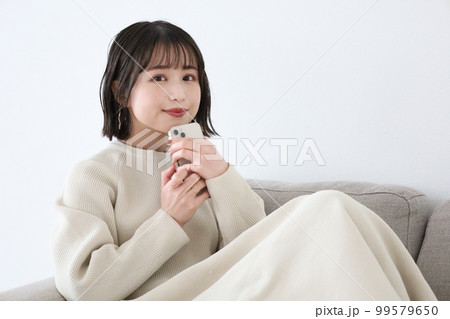 ソファーに座りスマホを持って微笑む女性 99579650