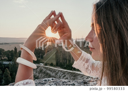 Happy woman in white boho dress making heart sign with hands on sunset in mountains. Romantic woman with long hair standing with her back on the sunset in nature in summer with open hands. 99580333