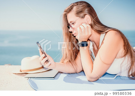 Digital nomad, woman in the hat, a business woman with a laptop sits on the rocks by the sea during sunset, makes a business transaction online from a distance. Freelance, remote work on vacation. 99580336