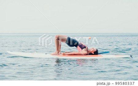 Woman sup yoga. Happy young sporty woman practising yoga pilates on paddle sup surfboard. Female stretching doing workout on sea water. Modern individual female outdoor summer sport activity. 99580337