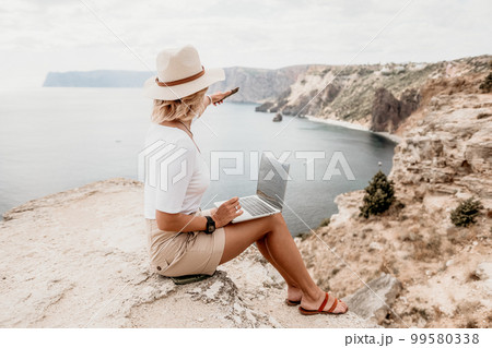 Digital nomad, Business woman working on laptop by the sea. Pretty lady typing on computer by the sea at sunset, makes a business transaction online from a distance. Freelance, remote work on vacation 99580338