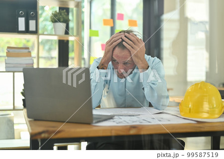 Worried architect man sitting at workplace holding his head on hands, feeling stressed and nervous Worried architect man sitting at workplace holding his head on hands, feeling stressed and nervous 99587519