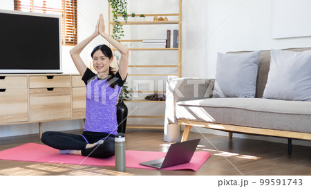 Beautiful healthy woman practicing yoga, Stretch or warm up before exercising, Calming the mind and meditating in her living room, Dumbbells and a protein shake or bottle of water, Healthy exercise. 99591743