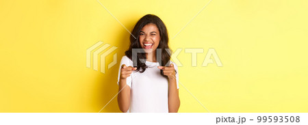 Portrait of cheeky, beautiful african-american woman in white t-shirt, winking and pointing fingers at camera to say congrats, praising you, standing over yellow background 99593508
