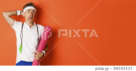 Concept of workout, gym and lifestyle. Image of awkward middle-aged athlete, looking confused, holding yoga mat and skipping rope, standing over orange background 99593525