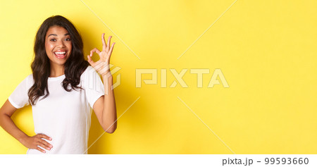 Portrait of happy good-looking african american girl, showing okay sign and smiling in approval, praise good work, standing over yellow background 99593660
