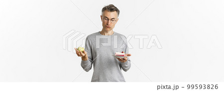 Portrait of thoughtful middle-aged man in glasses, making decision between tasty cake and green apple, standing over white background 99593826