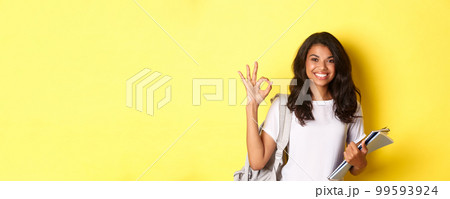 Portrait of satisfied african-american female student, smiling pleased and showing okay sign, like something good, standing over yellow background 99593924