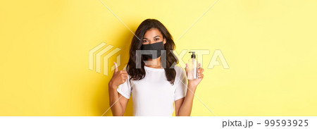 Concept of covid-19, social distancing and lifestyle. Portrait of cheerful african-american girl in face mask, recommend to use antiseptic, showing hand sanitizer and thumbs-up Concept of covid-19, social distancing and lifestyle. Portrait of cheerful african-american girl in face mask, recommend to use antiseptic, showing hand sanitizer and thumbs-up 99593925