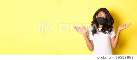 Concept of coronavirus, pandemic and lifestyle. Image of cute african-american girl in face mask, shrugging and looking clueless, dont know anything, standing over yellow background Concept of coronavirus, pandemic and lifestyle. Image of cute african-american girl in face mask, shrugging and looking clueless, dont know anything, standing over yellow background 99593998
