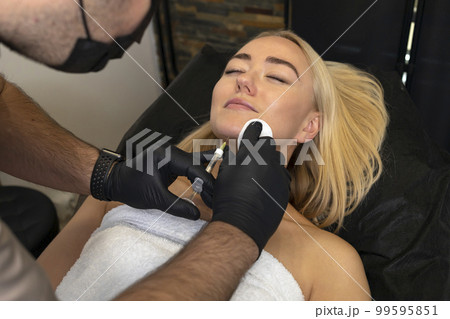Physician adds volume to woman's chin with filler, injection. White female lying on couch. Beauty physician holds syringe near girl's face.Chin reshaping, augmentation.Cosmetologist,aesthetic medicine 99595851
