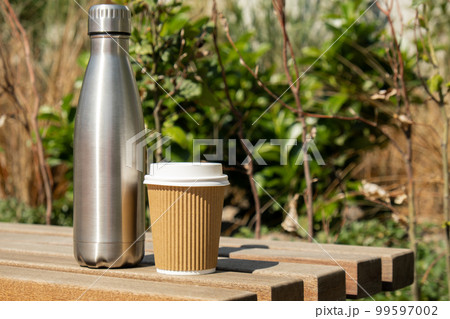 Reusable steel thermo water bottle on wooden bench with paper cup. Sustainable lifestyle. Plastic free zero waste free living. Go green Environment protection. Health-conscious Reusable steel thermo water bottle on wooden bench with paper cup. Sustainable lifestyle. Plastic free zero waste free living. Go green Environment protection. Health-conscious 99597002