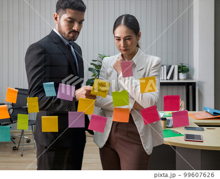 Businessman in black suit and colleague meeting with sticky paper notes on the glass wall for new ideas. Using agile methodology for business in a tech start-up office. Businessman in black suit and colleague meeting with sticky paper notes on the glass wall for new ideas. Using agile methodology for business in a tech start-up office. 99607626