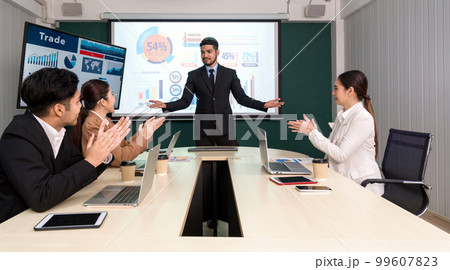 Businessman in black suit giving presentation with large digital monitor. Business executives team meeting in modern office with laptop computer, tablet, mobile phone and coffee on table. Businessman in black suit giving presentation with large digital monitor. Business executives team meeting in modern office with laptop computer, tablet, mobile phone and coffee on table. 99607823