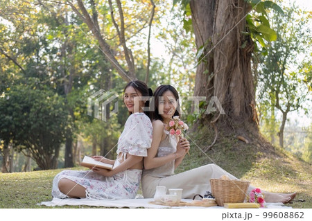 Charming beautiful young Asian woman in a lovely dress, holding a flower bouquet, enjoying a picnic with her friend in the park 99608862