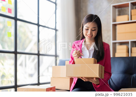 Young woman, an Asian online business owner, smiles, prepares parcel boxes and checks online orders for products to be delivered to customers 99621035