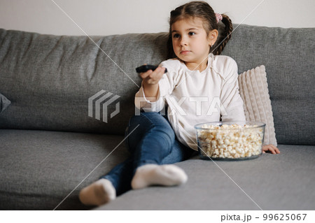 Cute little girl using remote controller of TV and watching cartoons. Four year girl eating popcorn on sofa Cute little girl using remote controller of TV and watching cartoons. Four year girl eating popcorn on sofa 99625067