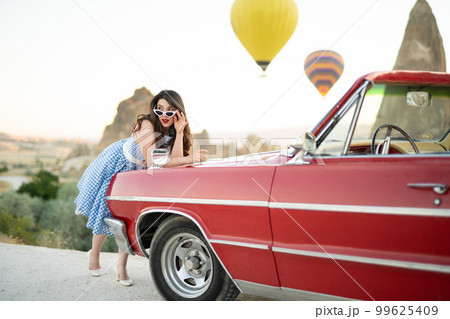 beautiful girl in retro style posing near a vintage red cabriolet car on background of balloons in Cappadocia. beautiful girl in retro style posing near a vintage red cabriolet car on background of balloons in Cappadocia. 99625409