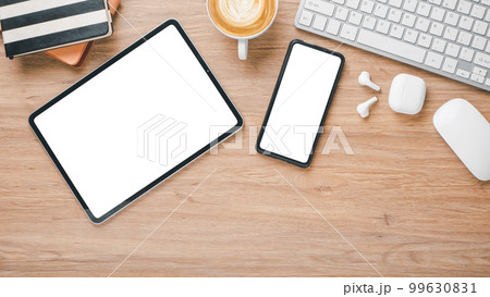 Wooden desk workplace with blank screen tablet and smartphone, mouse, earphone, notebook, keyboard and cup of coffee, Top view flat lay. 99630831