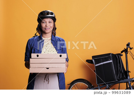 Portrait of deliverywoman wearing helmet and pizzeria uniform carrying pizza cardboxes during takeaway delivering. Asian restaurant employee smiling at camera in studio with yellow background Portrait of deliverywoman wearing helmet and pizzeria uniform carrying pizza cardboxes during takeaway delivering. Asian restaurant employee smiling at camera in studio with yellow background 99631089