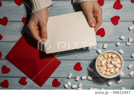Female hands holding empty valentine card mock up red envelope with white cup of coffee and marshmallows on wooden blue background. Romantic Small hearts Valentine day. Blank paper card copy space for Female hands holding empty valentine card mock up red envelope with white cup of coffee and marshmallows on wooden blue background. Romantic Small hearts Valentine day. Blank paper card copy space for 99632847