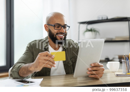 Online shopping. Excited latin businessman using tablet and credit card, making payment via bank app, sitting at desk Online shopping. Excited latin businessman using tablet and credit card, making payment via bank app, sitting at desk 99637358