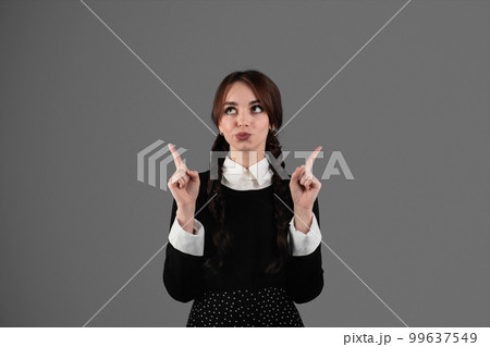 Pensive glad young girl student in gothic black clothes with pigtails shows fingers up to free space Pensive glad young girl student in gothic black clothes with pigtails shows fingers up to free space 99637549