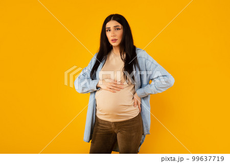 Sad young pregnant woman having back pain, touching belly and ache zone, suffering from backache, yellow background Sad young pregnant woman having back pain, touching belly and ache zone, suffering from backache, yellow background 99637719
