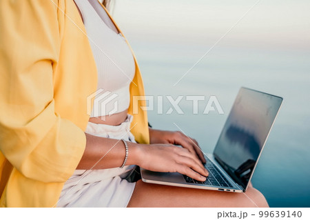 Digital nomad, Business woman working on laptop by the sea. Pretty lady typing on computer by the sea at sunset, makes a business transaction online from a distance. Freelance, remote work on vacation 99639140